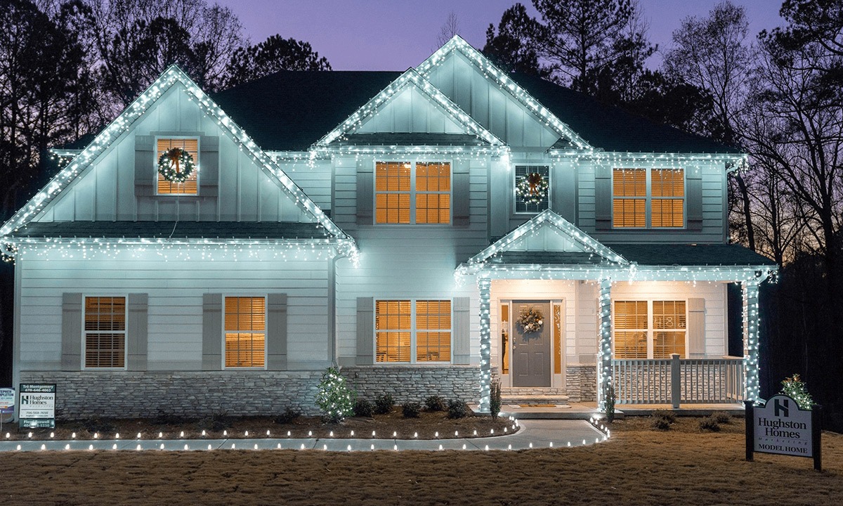 A two-story home decorated with cool white Christmas lights outlining the roofline, windows, and porch. Icicle lights hang from the eaves, and a pathway lined with glowing bulbs leads to the front door. Holiday wreaths on the windows and door complete the festive display. | Icy Lights LLC