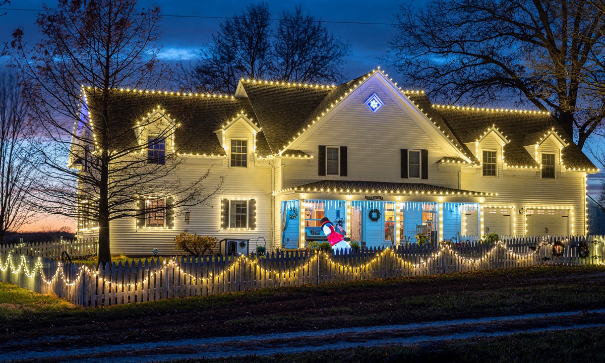 A two-story white house beautifully decorated with warm white Christmas lights outlining the roofline, dormer windows, and porch. Icicle lights hang from the porch roof, and the white picket fence is draped with glowing light garlands. A lit inflatable Santa Claus adds a festive touch in front of the porch. Bare trees surround the home, with a dark blue evening sky in the background | Icy Lights LLC